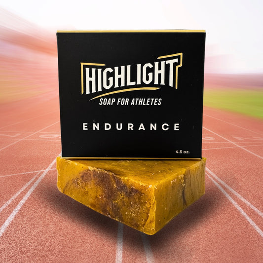 A black box of Highlight Soap for Athletes, labeled 'Endurance,' is perched on a rugged, amber-colored soap bar. The background features a blurred running track, conveying a sense of speed and stamina, perfectly aligning with the soap's theme of endurance.